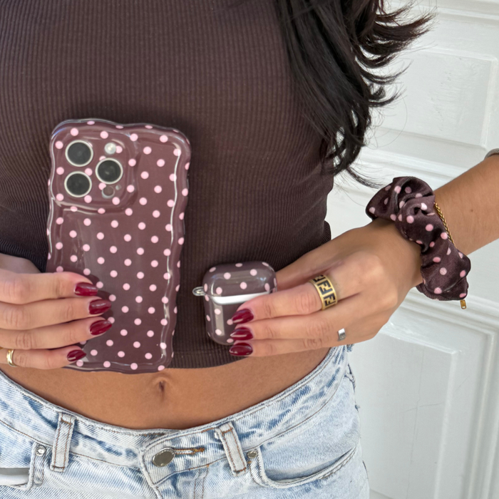 Woman holding a brown polka dot Coconut Lane AirPods case and matching scrunchie, wearing a casual outfit.
