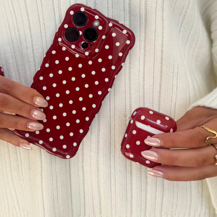 Hands holding a burgundy polka dot AirPods case and red polka dot phone case, wearing a white ribbed sweater.
