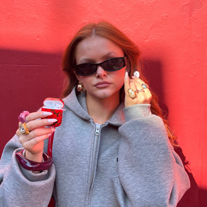 Woman in gray hoodie with sunglasses holding a Coconut Lane Leopard Hibiscus Airpods Case by a red wall.