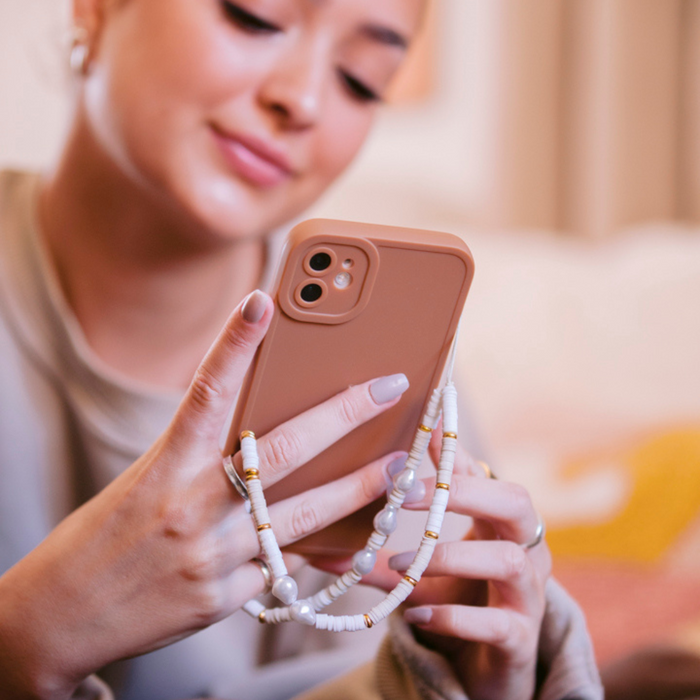 A person holds a smartphone adorned with Coconut Lane's Beaded Phone Strap in Nude & Pearl and a beige phone case. They are smiling while sitting indoors, their nails manicured in a light color. The background is softly blurred, contributing to the warm and cozy atmosphere.