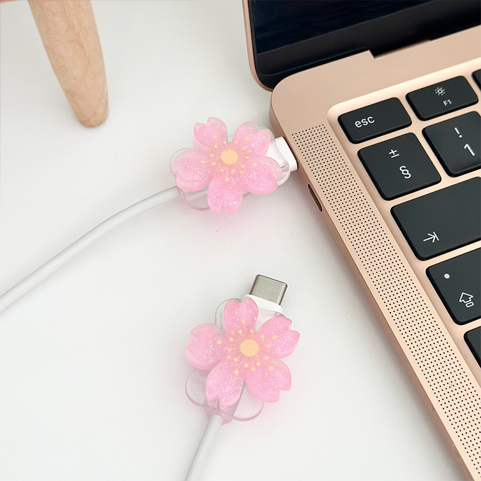Bright pink flower Coconut Lane cable cover charm on a white laptop charging cable, resting on a white surface.