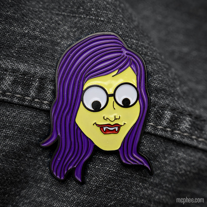 The Monster Girl Strange Pin showcases a cartoon character with purple hair, glasses, googly eyes, and fangs set against a dark fabric background. Made from diecast metal, it captures attention effortlessly.