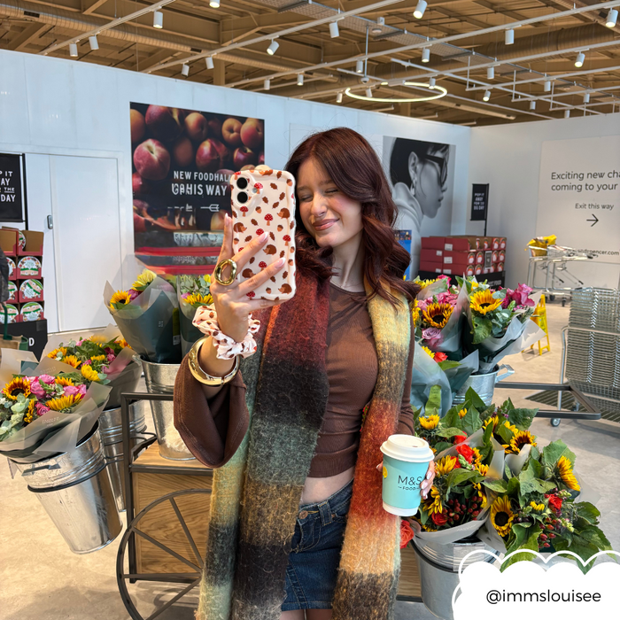 Smiling woman in-store holds coffee and a wavy green phone case with a forest animal pattern, standing by flower bouquets.