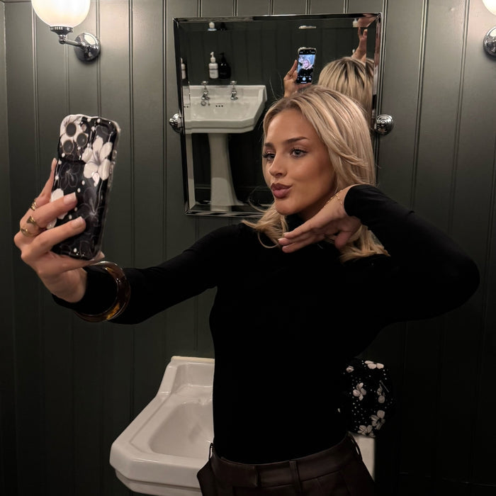 Woman in black takes a mirror selfie in a bathroom, holding a phone with a wavy black and white floral patterned case.