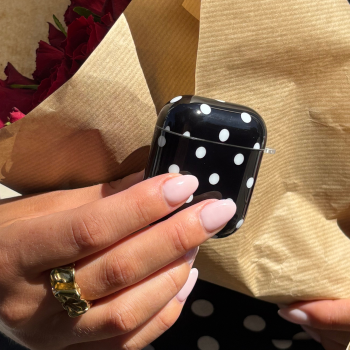 Manicured hand holding a black polka dot AirPods case and a brown paper-wrapped bouquet.