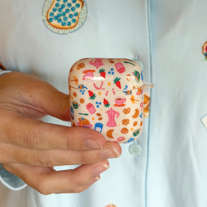Hand holds Coconut Lane Breakfast Club AirPods case with pastel design; person wears light blue embroidered pajamas.