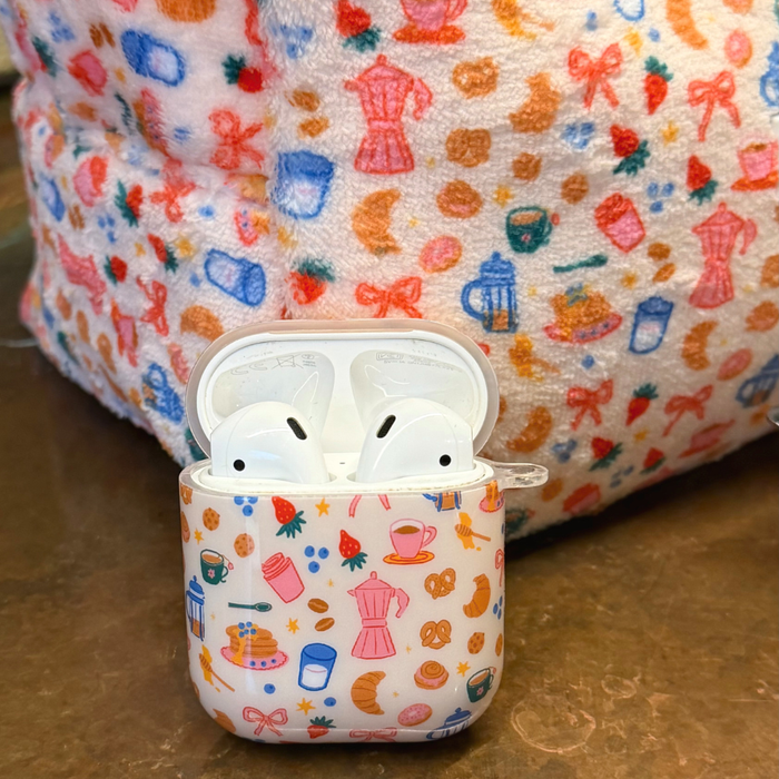 Breakfast Club AirPods case with colorful breakfast-themed pattern, shown with matching coffee and pastry print fabric behind.