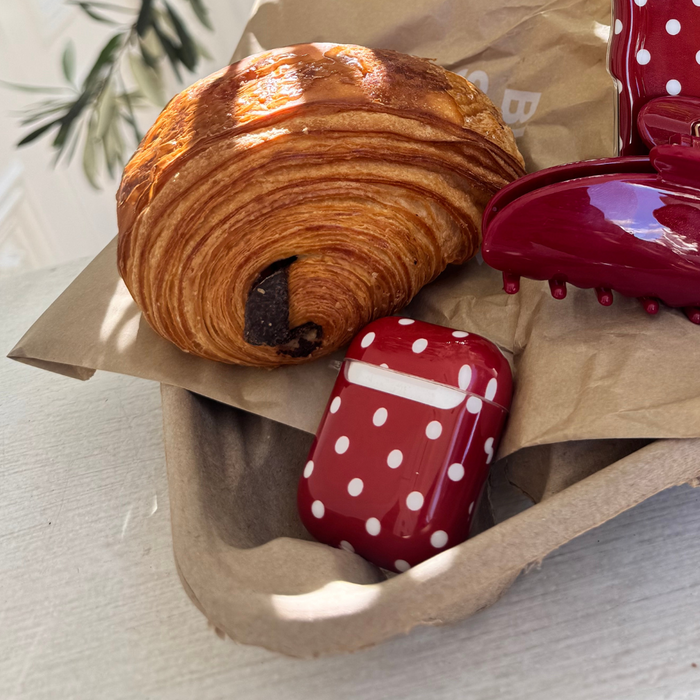 Burgundy polka dot AirPods case and matching hair clip beside a chocolate croissant on brown paper.
