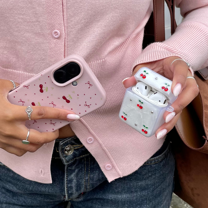 Pink sweatered person holds a phone and glossy white AirPods case, both decorated with red cherry patterns.