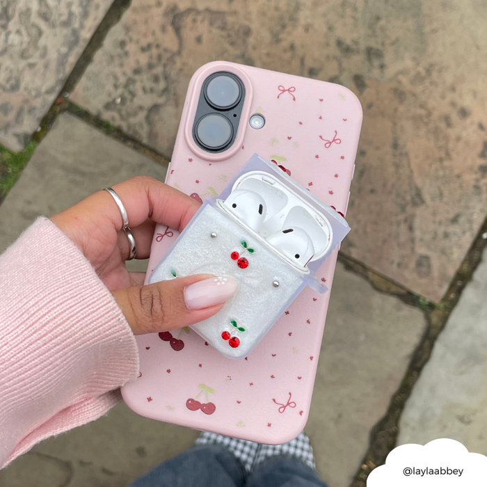 Hand holding a pink phone and Coconut Lane Airpods Case in Cherry Pearl, featuring cherry and pearl decorations.