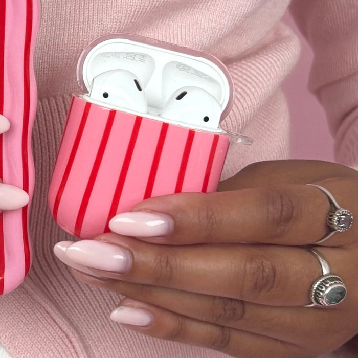 Hand with pale pink nails holds a Coconut Lane AirPods Case with cherry and stripe pattern; silver rings and pink sweater shown.