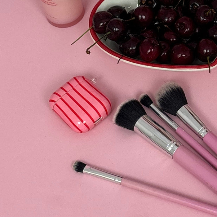 Pink makeup brushes and Cherry Stripes AirPods case on a pink surface with scattered cherries and a small bottle.