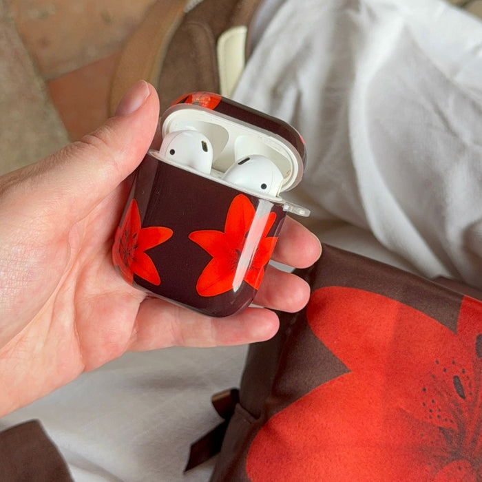 A hand holds a Coconut Lane Airpods Case in Cinnamon Lily, featuring a red floral pattern.