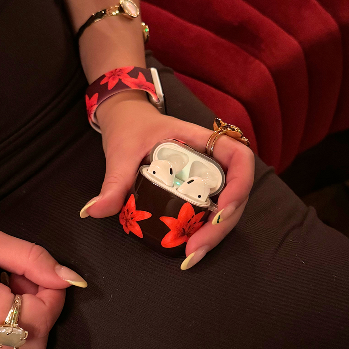 Hand holds a cinnamon-colored Coconut Lane Airpods case, with floral wristband, rings, and a black outfit visible.