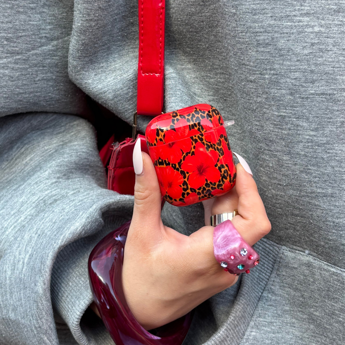 A hand with white nails holds a Coconut Lane AirPods case featuring a leopard and hibiscus pattern, shown against a gray hoodie.
