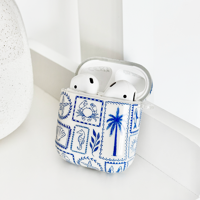 White wireless earbuds in a Malibu Drive AirPods case with coastal design, TPU cover, resting on a white shelf.