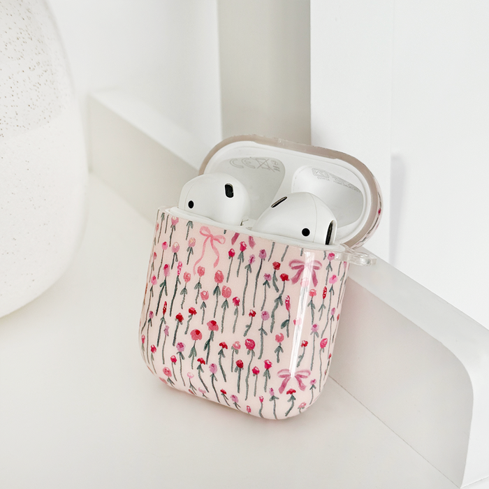 White wireless earbuds in a pink Peony Bow Coconut Lane AirPods case on a white surface next to a round textured object.
