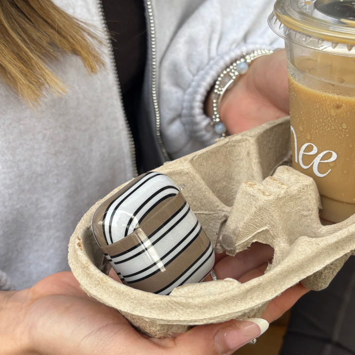 Hand holding a coffee cup and Coconut Lane Airpods Case with retro striped pattern, grey jacket, and beaded bracelet visible.