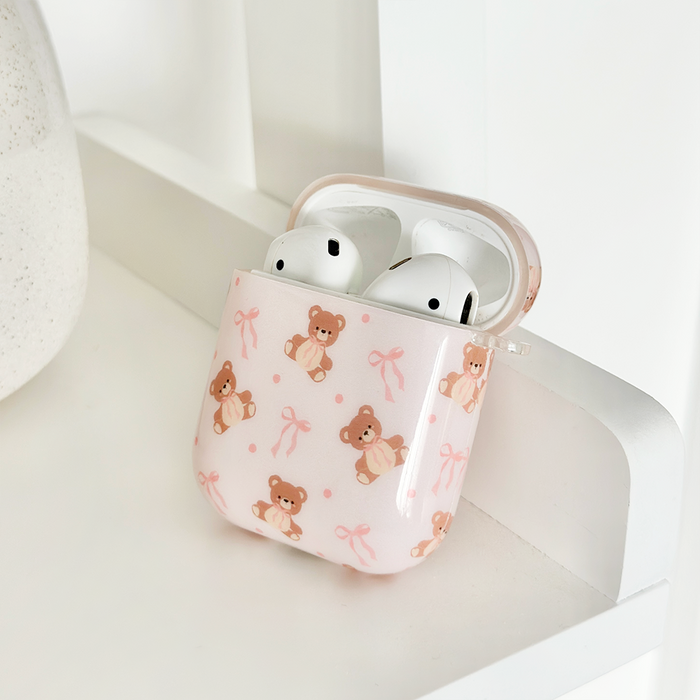AirPods in a Coconut Lane Thea Teddy case featuring pink bows and teddy bears, resting on a white shelf.