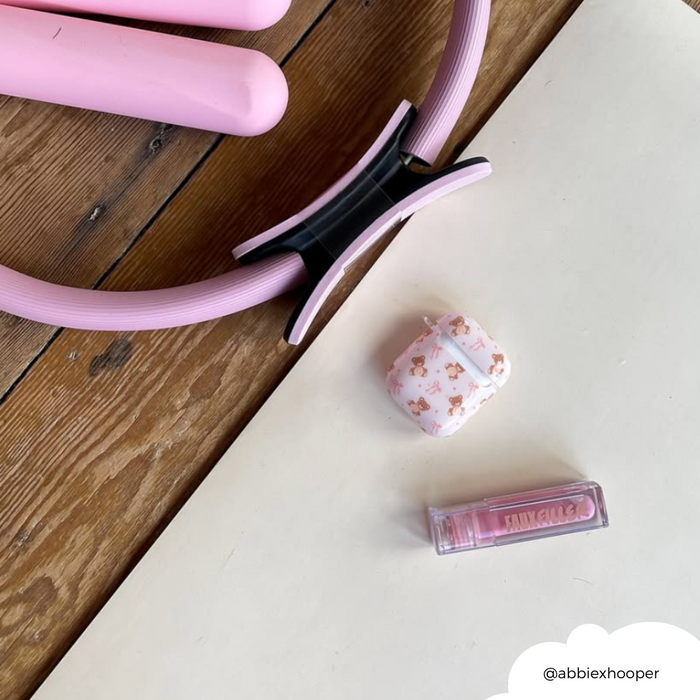 Pink exercise ring, fuzzy beige AirPods case, and pink lip gloss on a wooden and white surface.
