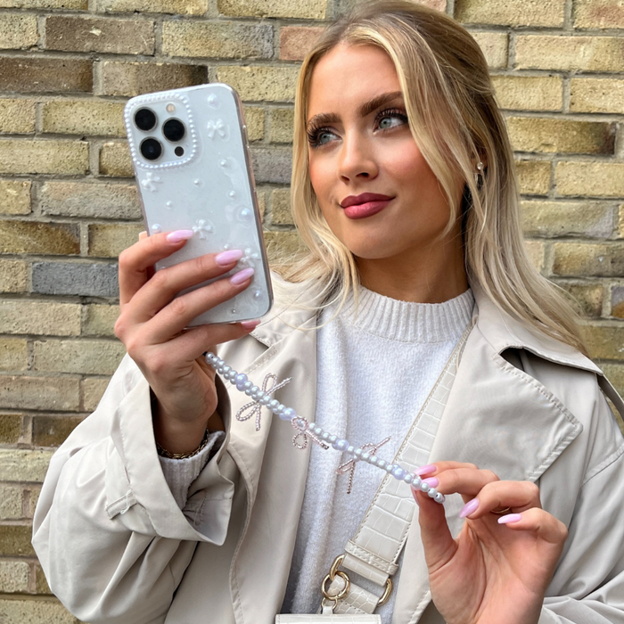 Blonde woman taking a selfie with a white phone, showing a beaded phone strap with small pearl bows.