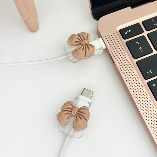 Chargers with nude bow cable cover charms beside a rose gold laptop on a white surface.