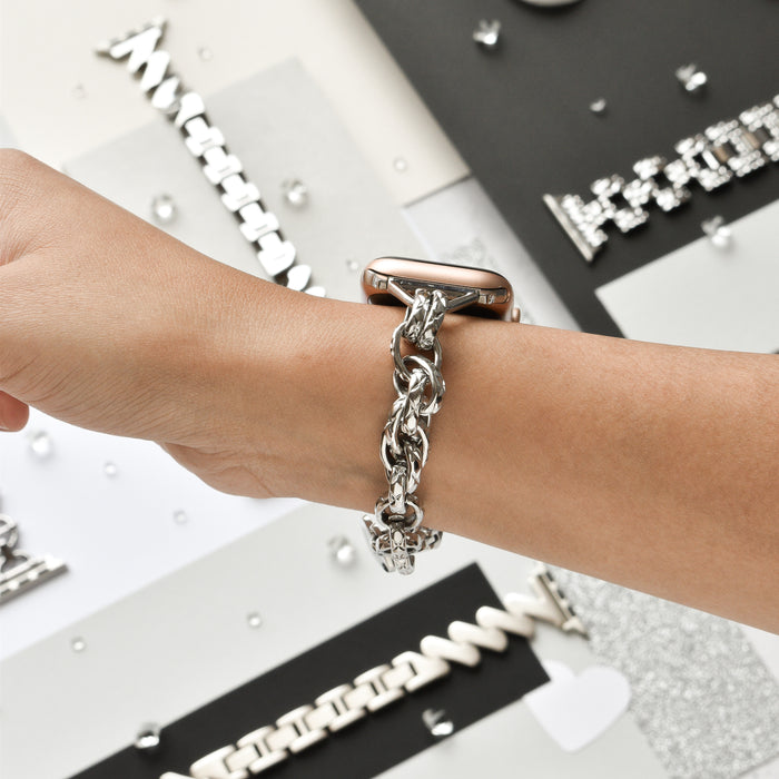 A person wearing the Coconut Lane Chain Bracelet Apple Watch Strap - Silver on their wrist. The background reveals various silver jewelry pieces and accessories displayed on different textured surfaces, including an elegant silver chain bracelet.