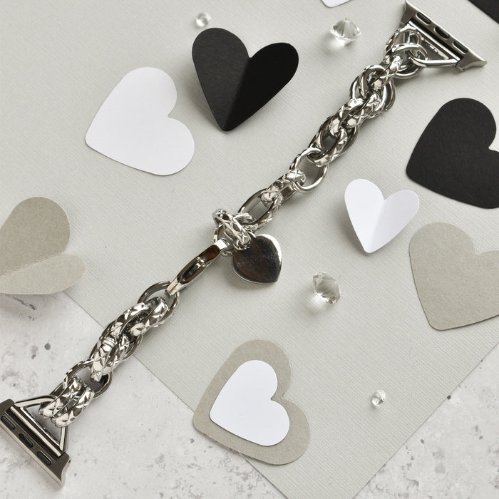 A Chain Bracelet Apple Watch Strap in silver, by Coconut Lane, rests on a textured surface adorned with black, white, gray, and silver paper hearts. Small transparent crystals are scattered around, adding a touch of sparkle to the composition. The scene is reminiscent of an elegantly crafted bracelet with a heart charm.