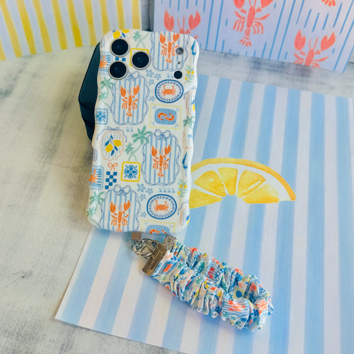 A Coastal Catch scrunchie phone wrist strap featuring a blue tile, orange lobster, and yellow lemon pattern attached to a matching Wavy Luxe phone case on a blue and white striped background.