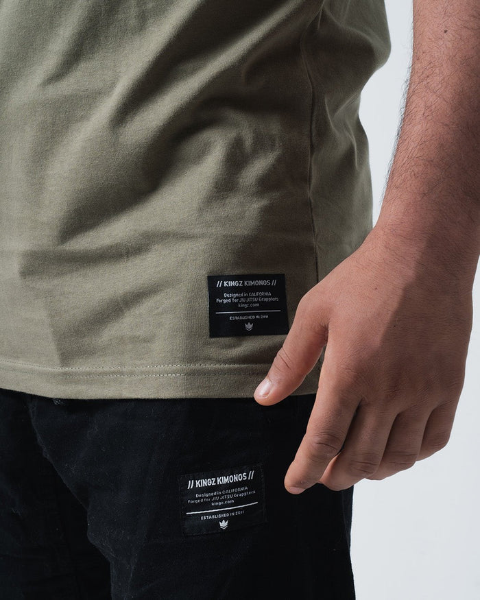Kingz Est. 2011 Tee Military Green - Close-Up Side View Male Hand Black Label Text Bottom Hem Streetwear BJJ Jiu-Jitsu Casual Apparel [color:Military Green]