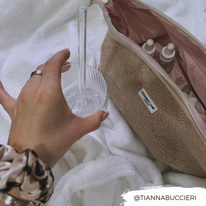 Coconut Lane Teddy Rupert expandable beige makeup bag and skincare bottles on a white towel, hand holding a glass nearby.