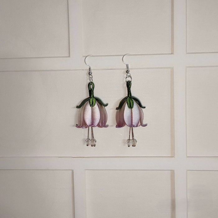 A pair of handmade Chanhua flower dangle earrings on a white textured background. The earrings feature light purple petals and a green stem, with small crystal beads at the bottom.