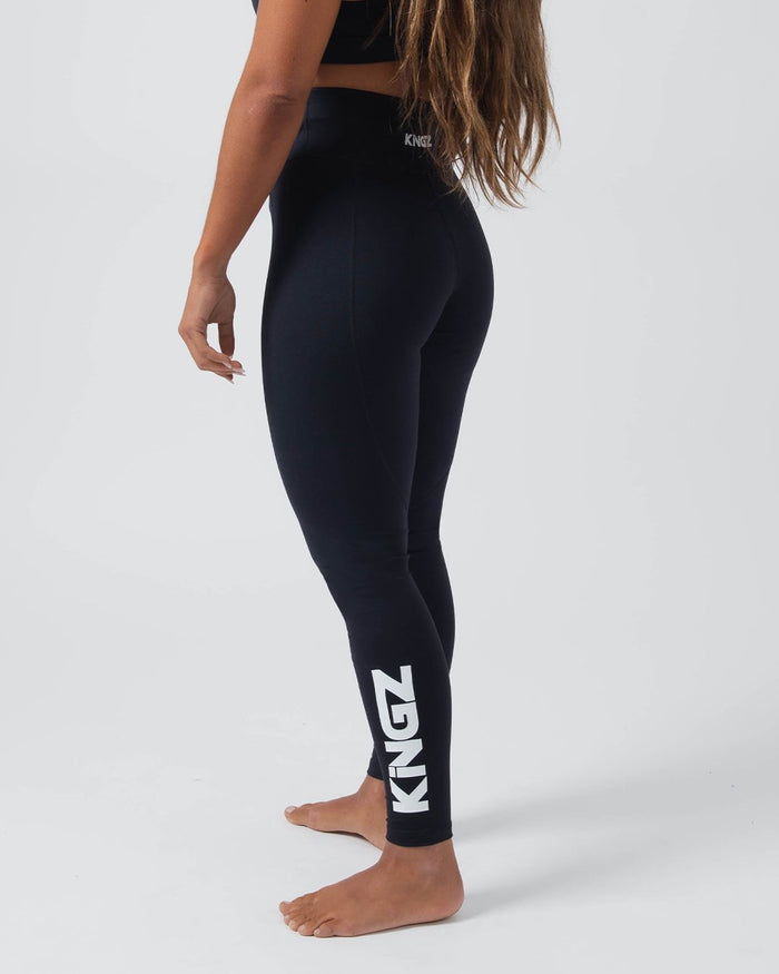 Kingz Kore Women's Grappling Spats Black - Full Body Left Side Profile Female Athlete All-Black KINGZ Large White Text Waistband KINGZ White Vertical Text Right Calf High-Waist Women's No-Gi BJJ Spats Kore Collection [color:Black]