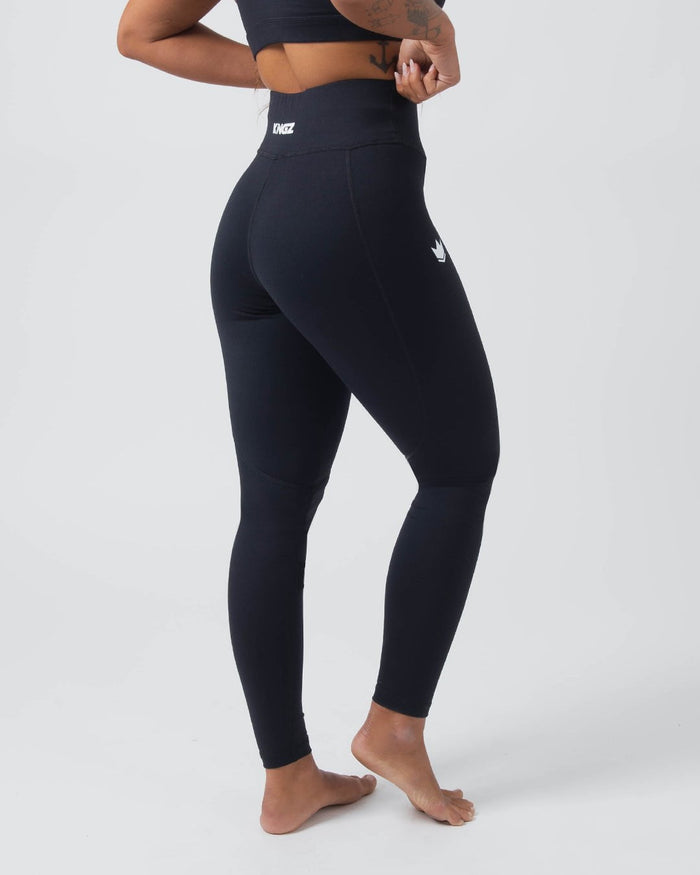Kingz Kore Women's Grappling Spats Black - Full Body 3/4 Angled Front View Female Athlete All-Black Small White Crown Logo Left Thigh KINGZ White Waistband Text Hands Behind Back Pose Women's No-Gi BJJ Spats Kore Collection [color:Black]