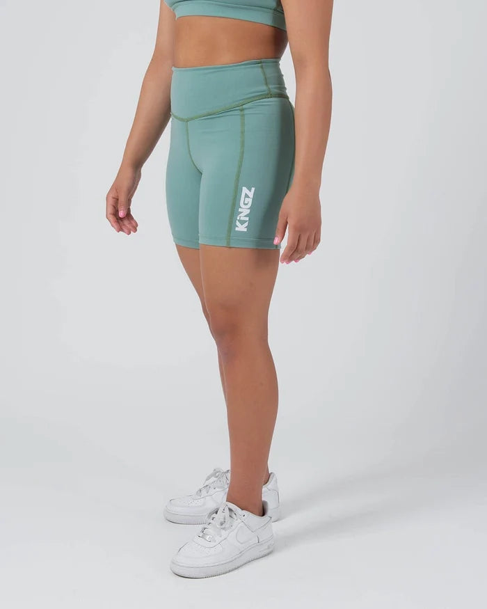 Kingz Kore Women's Training Shorts Green Light Sage - Full Body Left Side Profile Female Athlete Sage Mint Green Biker Style Short Cut High-Waist KINGZ White Vertical Text Right Outer Thigh Olive Green Contrast Side Seam Stitching Matching Sage Sports Bra White Sneakers Women's Training Shorts No-Gi BJJ Kore Collection [color:Light Green]