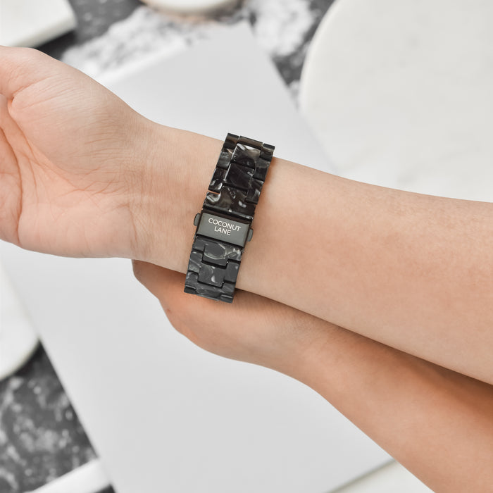 A close-up of a person's wrist wearing a Luxe Black Marble Apple Watch Strap with a glossy finish, branded with the text "Coconut Lane." The person’s other hand appears to be adjusting the strap. The background includes a marble-patterned surface.