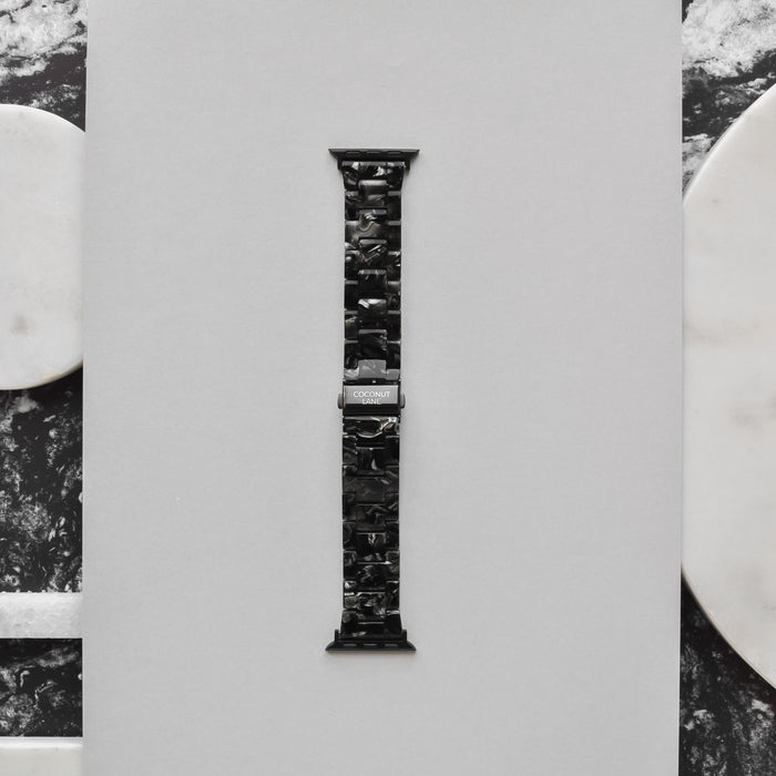 A Luxe Black Marble Apple Watch strap from Coconut Lane is displayed vertically against a light gray background. The band features a marbled pattern and a shiny metal clasp, with the brand name "Coconut Lane" engraved on it. The background includes marble accents in white and black on the sides.