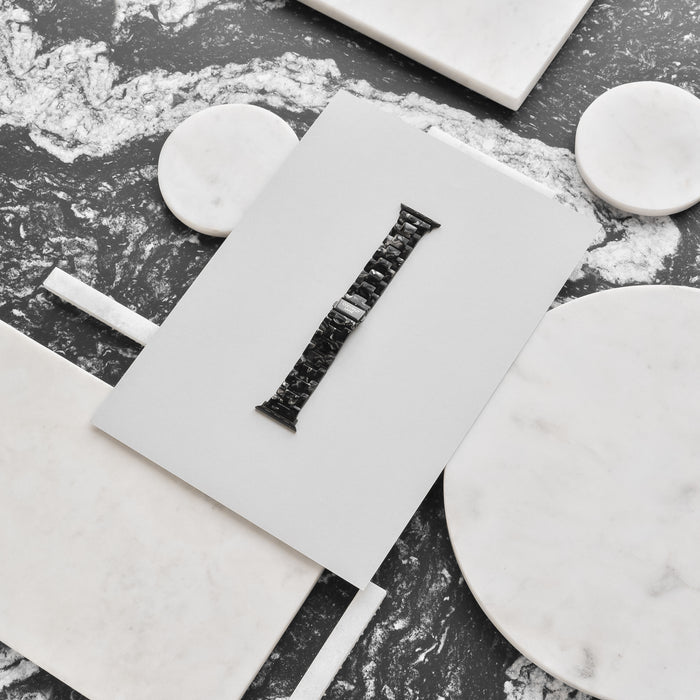 A Luxe Black Marble Apple Watch Strap from Coconut Lane is displayed on a white rectangular card, laid on a marble surface with various round and rectangular marble pieces around it. The watch strap is centered and stands out against the contrasting white and grey marbled background.