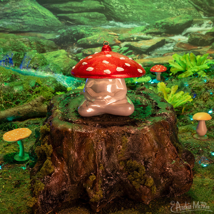 A Meditating Mushroom Ornament is perched on a tree stump, enveloped by the verdant foliage of the forest, creating a peaceful ambience that invites contemplation and serenity amidst nature.