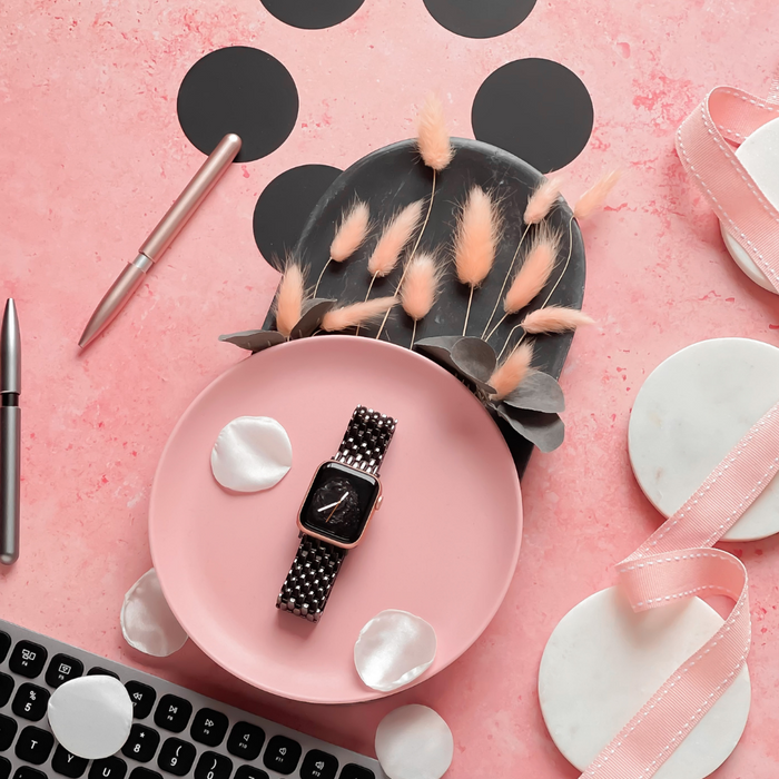 A flat lay on a pink textured surface featuring the Coconut Lane Mermaid Apple Watch Strap in black with a checkered band on a pink plate, surrounded by white petals, a corner of a black keyboard, a silver pen, and decorative dried flowers in a gray vase. Ribbons and marble coasters are also present.