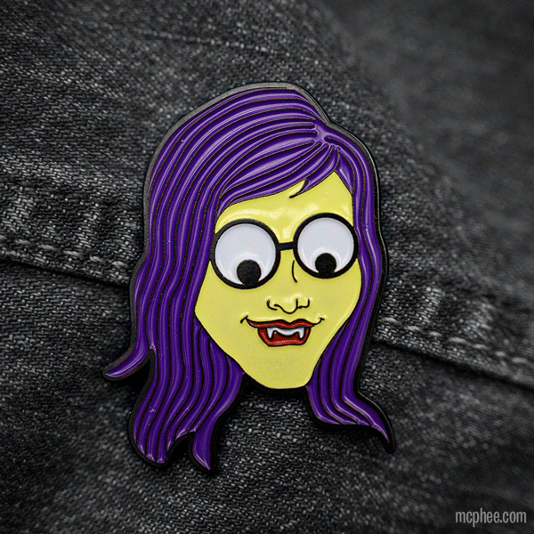 The Monster Girl Strange Pin, featuring a cartoonish vampire face with purple hair and glasses, makes for an eye-catching accessory on denim fabric. Its playful design and quality craftsmanship add charm to any outfit.