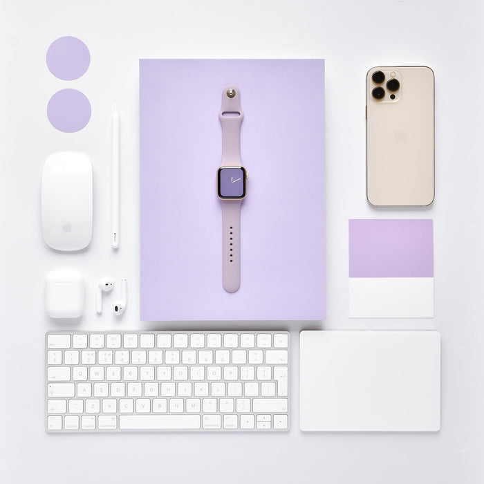 A neatly arranged flat lay of tech gadgets and accessories, including a lavender smartwatch with the NAKD Apple Watch Strap - Lavender Lilac from Coconut Lane, a silver smartphone, a wireless mouse, a stylus, earbuds, a keyboard, and a touchpad, all on a white and lavender background.