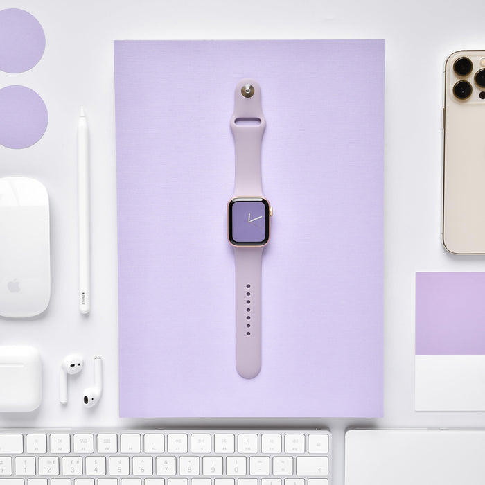 A purple smartwatch with the NAKD Apple Watch Strap in Lavender Lilac, from Coconut Lane, is centered on top of a lavender-colored sheet. Surrounding it are various Apple products including an iPhone, AirPods, Apple Pencil, mouse, and keyboard, all arranged on a white background.