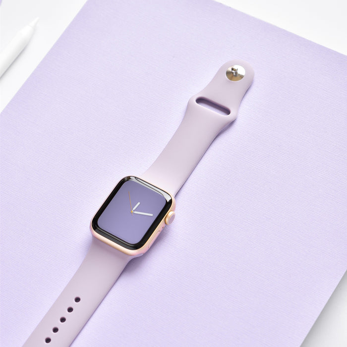 A Coconut Lane smartwatch with a gold frame and NAKD Apple Watch Strap in Lavender Lilac is displayed against a lavender-colored background. The minimalist clock face on the screen complements its sleek design. A white stylus pen from the NAKD Collection is partially visible on the left side of the image.