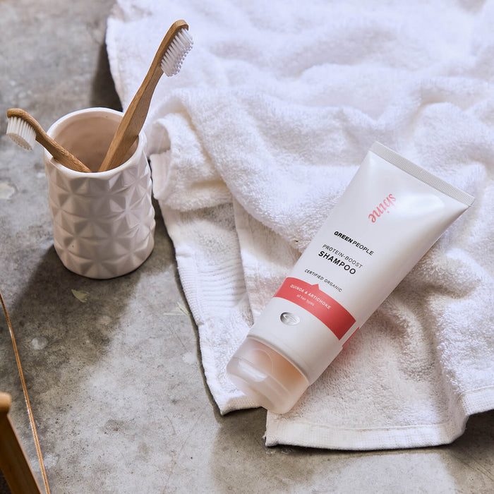 Protein-boost shampoo lying on a white towel on stone countertop with natural wooden toothbrushes in a cup.