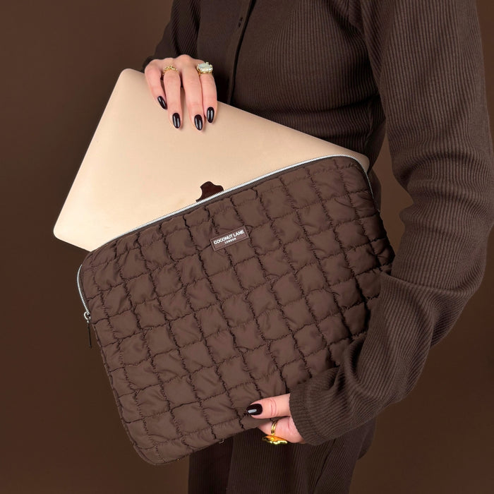 Person in brown holds a gold laptop and a Coconut Lane quilted chocolate laptop sleeve with a logo patch.
