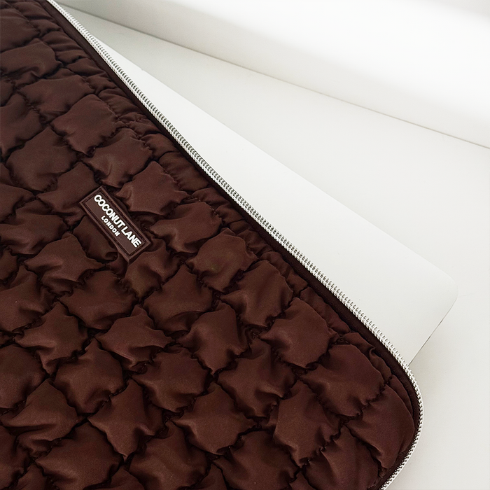 Silver laptop partially inside a brown quilted Coconut Lane laptop sleeve, resting on a white surface.