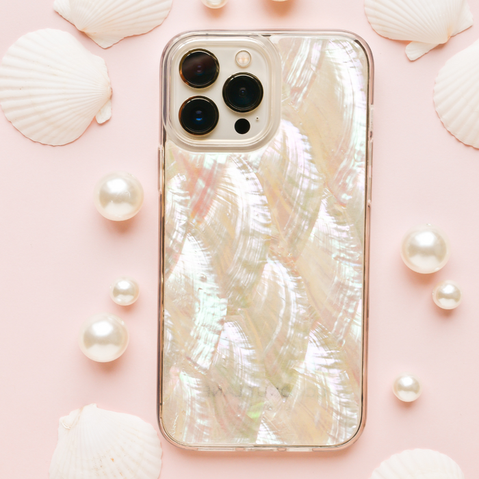 A smartphone adorned with Coconut Lane's Real Natural Shell Phone Case - White rests on a pastel pink background, surrounded by scattered white seashells and pearls, creating a coastal, elegant aesthetic.
