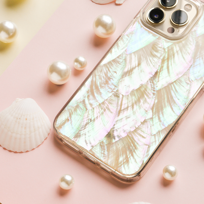 A smartphone with a shimmering, iridescent case designed to resemble fish scales lies on a pink and yellow background. The scene, featuring the Real Natural Shell Phone Case in White by Coconut Lane, is decorated with pearls and small white seashells, creating a delicate, ocean-themed aesthetic.