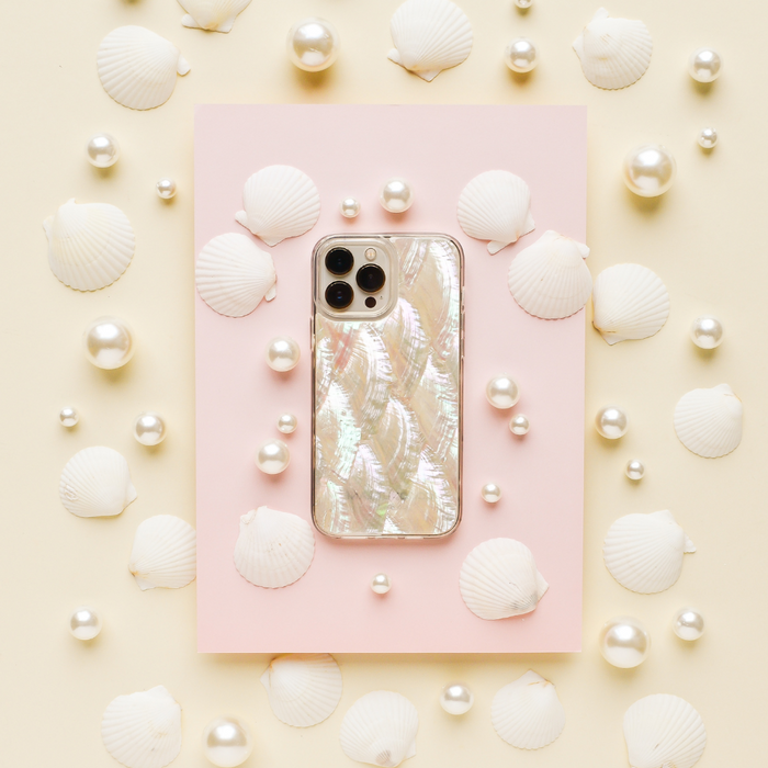 A white iPhone adorned with Coconut Lane's Real Natural Shell Phone Case is set on a pastel pink rectangular card, surrounded by white seashells and pearls, all against a cream-colored background. This arrangement creates a marine-themed aesthetic with a soft, elegant feel.